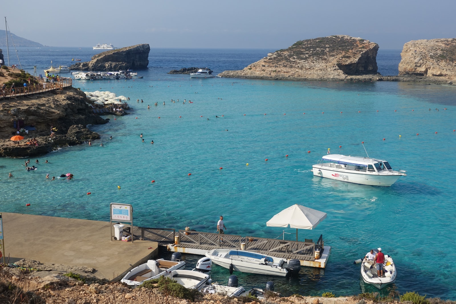 Swimmers in the Blue Lagoon with a busy jetty, boats, and umbrellas visible on the rocky shoreline.