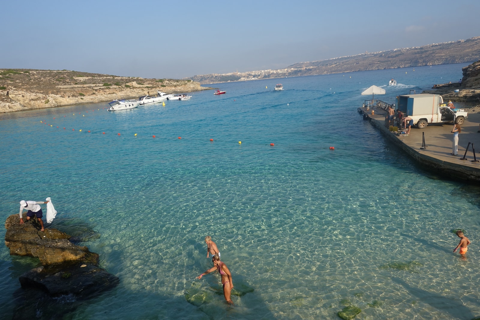 A wide view of the Blue Lagoon, showing swimmers in the clear turquoise water with boats and the rocky shoreline.