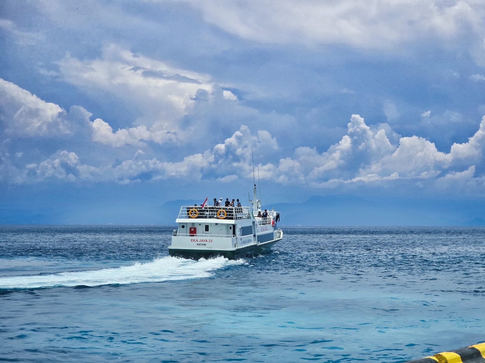 A passenger boat sails on the water with the green hills of Nusa Penida visible in the background under a blue sky.