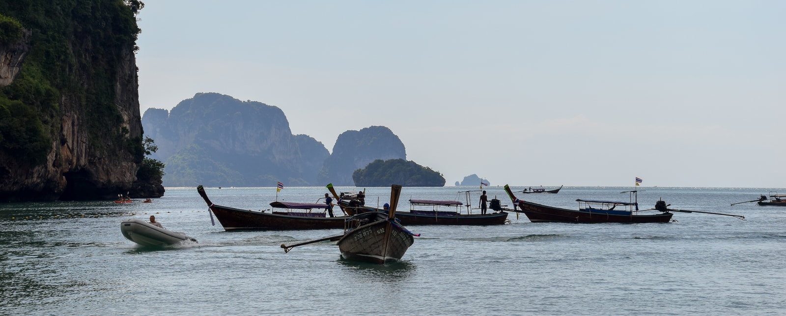 A fleet of traditional longtail boats waits in the shallow, clear waters near the shore of the Railay Peninsula, with dramatic cliffs in the background.