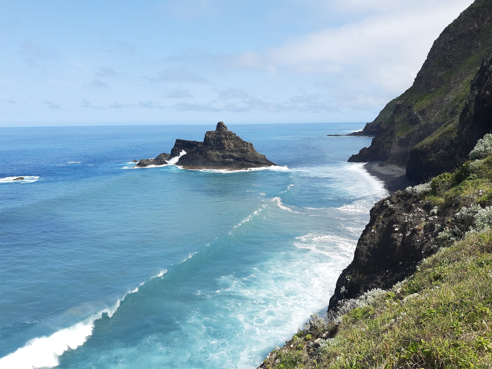 A dramatic aerial view of a rugged green coastline with a small sea stack and turquoise water.