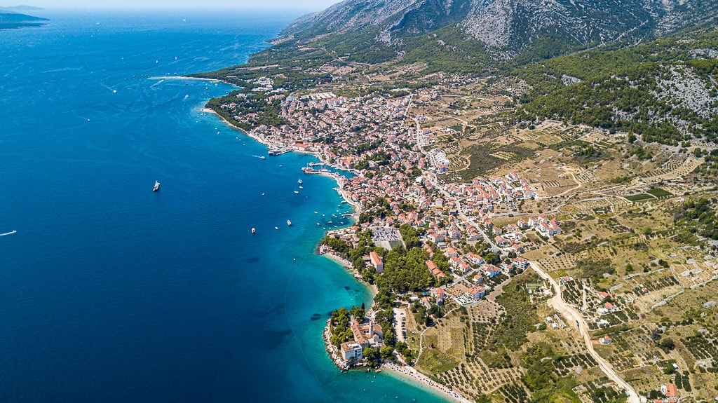 An aerial view shows the harbor town of Bol, with its red-roofed buildings clustered along the coast beneath a dramatic mountain ridge.