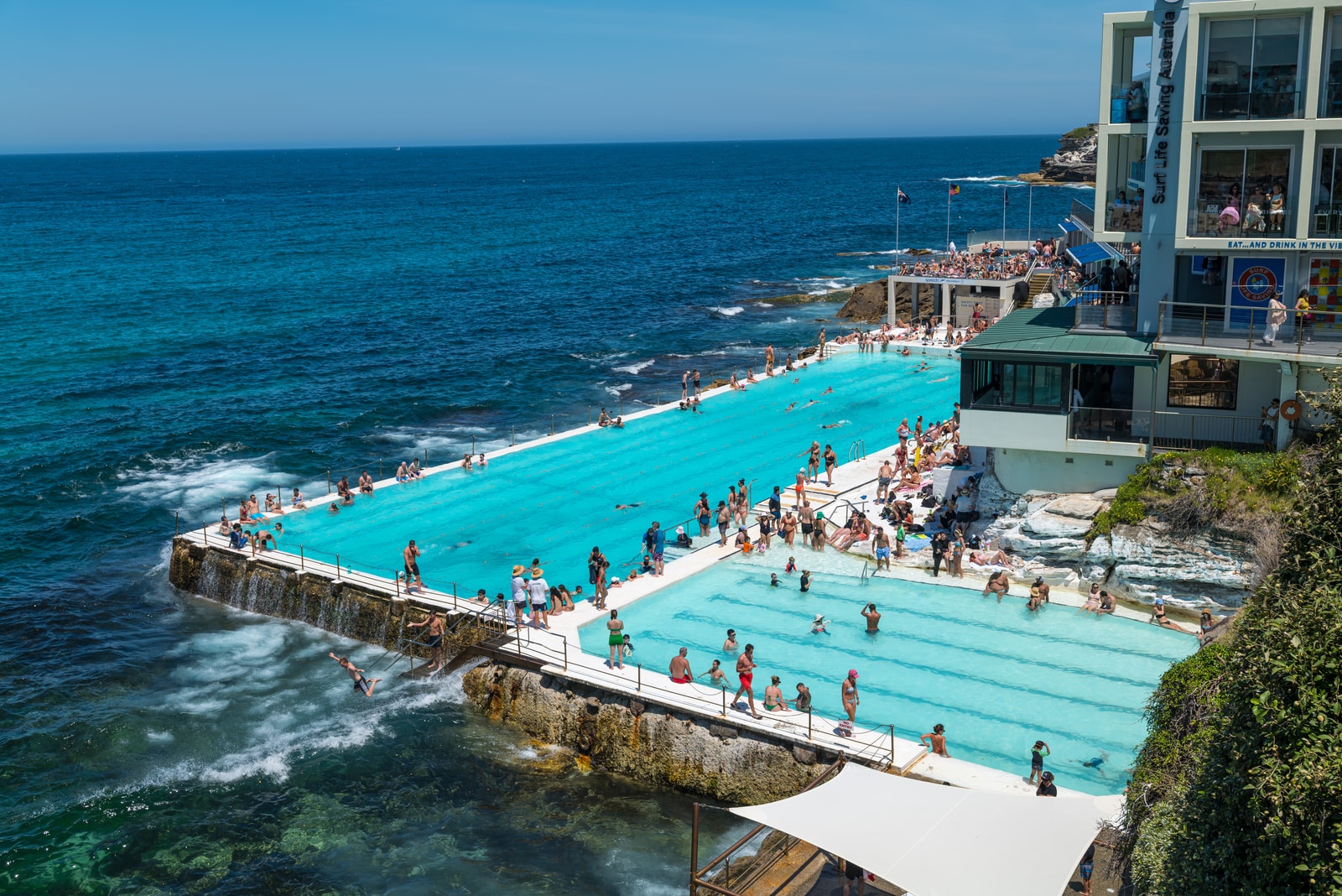 The bright turquoise water of the Bondi Icebergs ocean pool is set against the deep blue of the Pacific Ocean, with waves crashing over the edge.