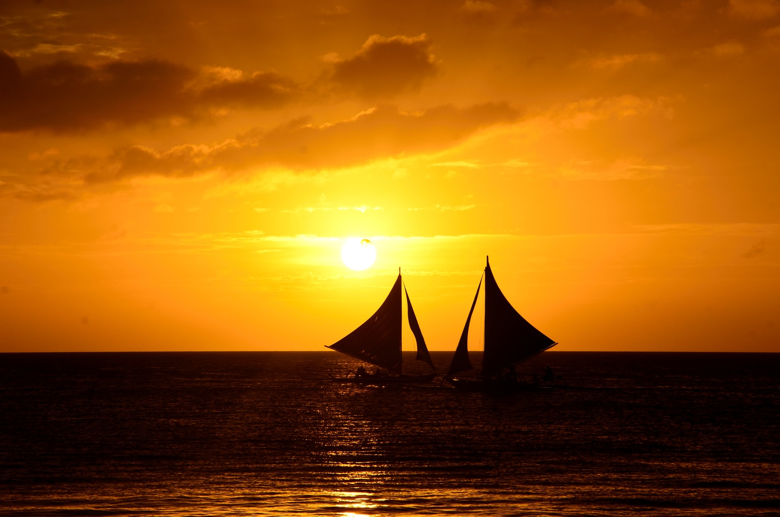 Silhouetted paraw sailboats cross the water at sunset, which casts a golden glow on the sea.