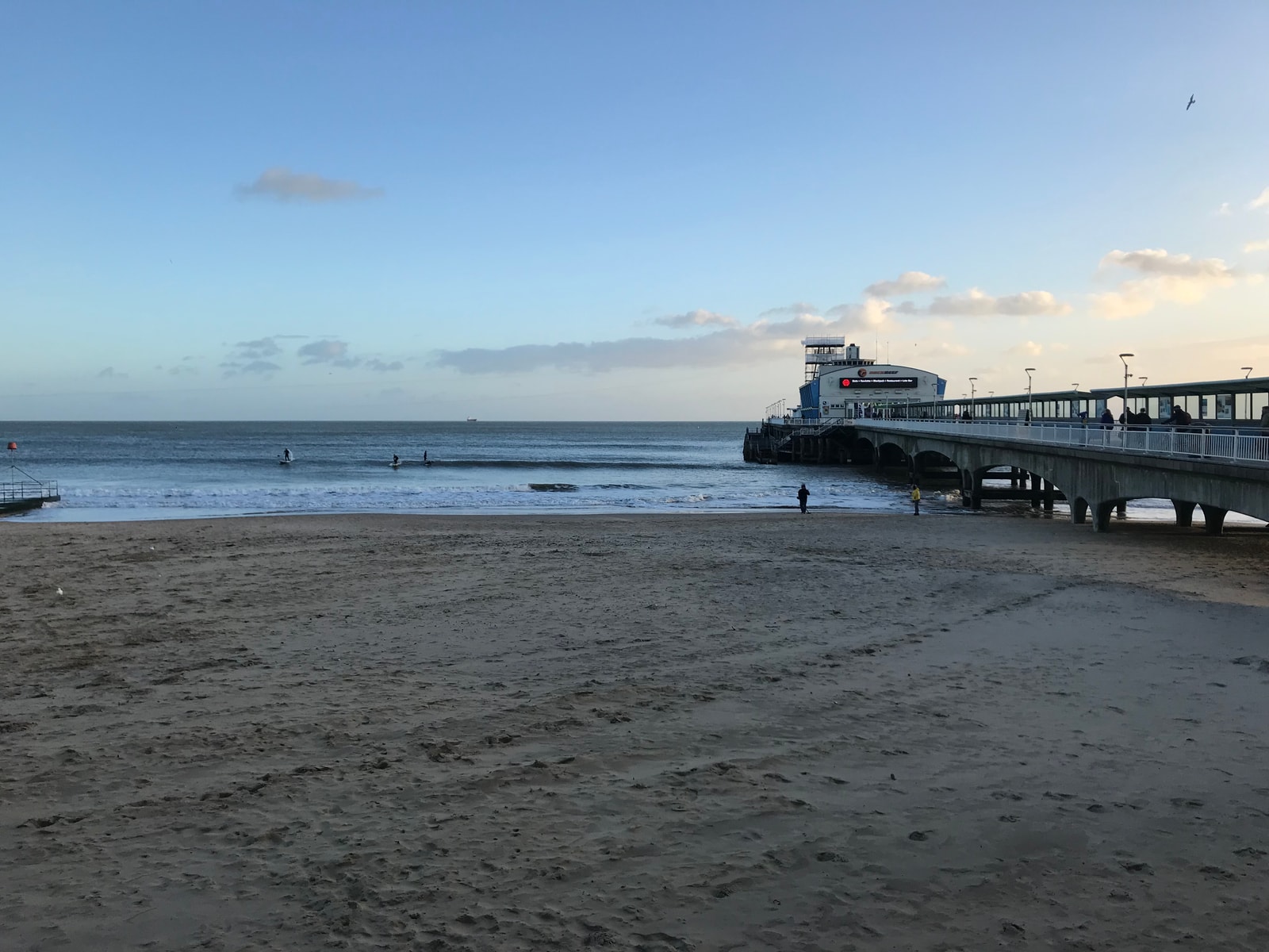 A wide-angle daytime shot shows Bournemouth Pier extending into the blue sea under a clear sky, with the bustling central beach in the foreground.