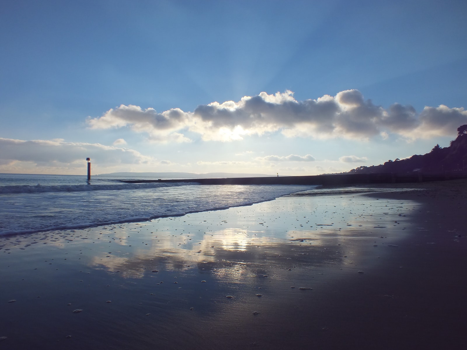 Sun rays break through clouds at sunset, reflecting on the wet sand and a wooden groyne at Bournemouth Beach.