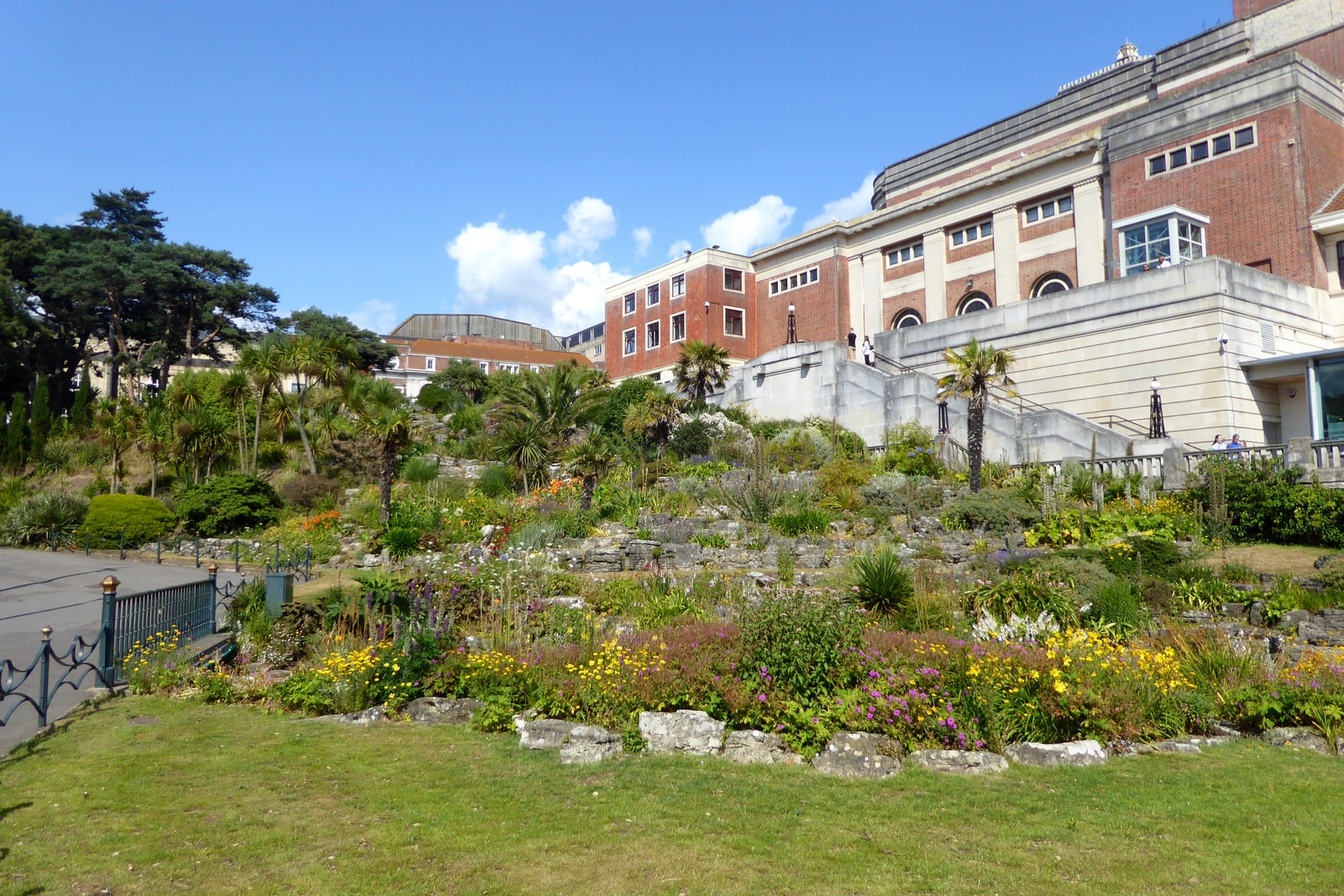 The terraced rockery of Bournemouth's Lower Gardens is filled with colorful flowers on a sunny day, with the historic Pavilion building in the background.