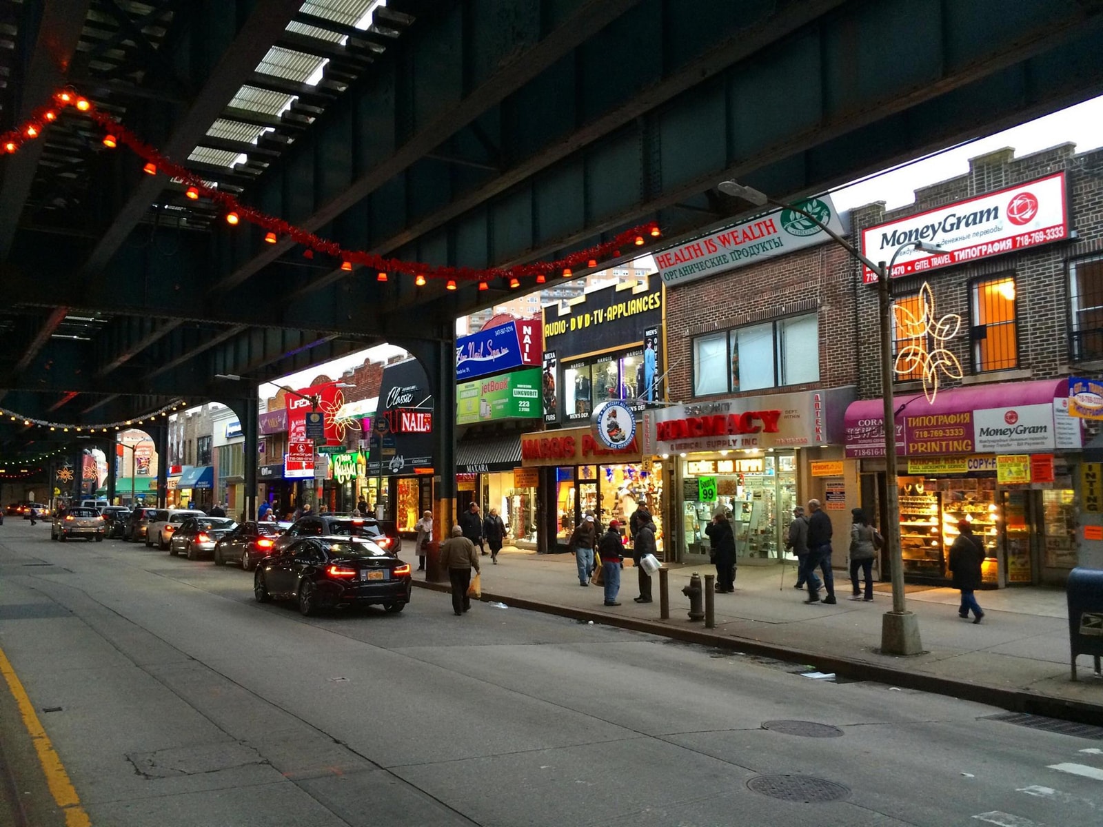 A bustling street scene on Brighton Beach Avenue, directly underneath the dark metal structure of the elevated subway tracks.