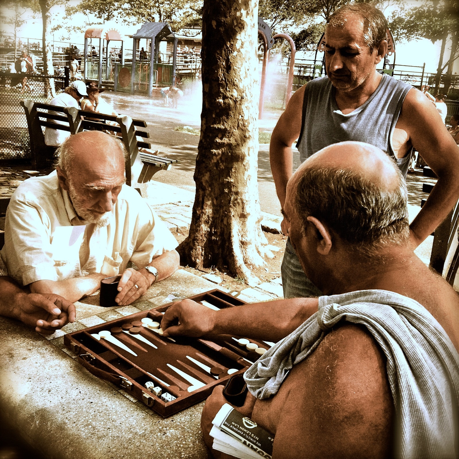A candid photo of older men gathered around a table outdoors, deeply focused on a game of backgammon.