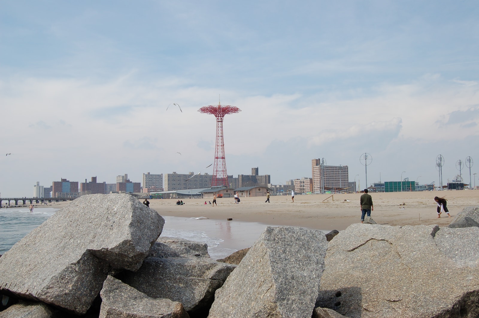 A wide view of the sandy shoreline at Brighton Beach, with the iconic Parachute Jump tower of Coney Island visible in the distance.