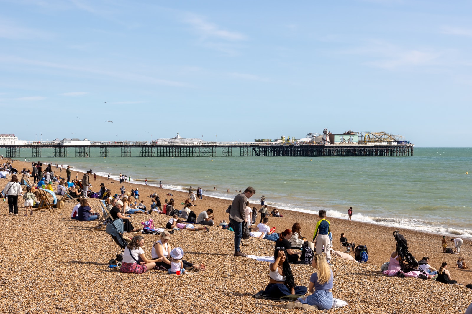 People enjoying the pebble beach in Brighton, with the iconic Palace Pier in the background under a blue sky.