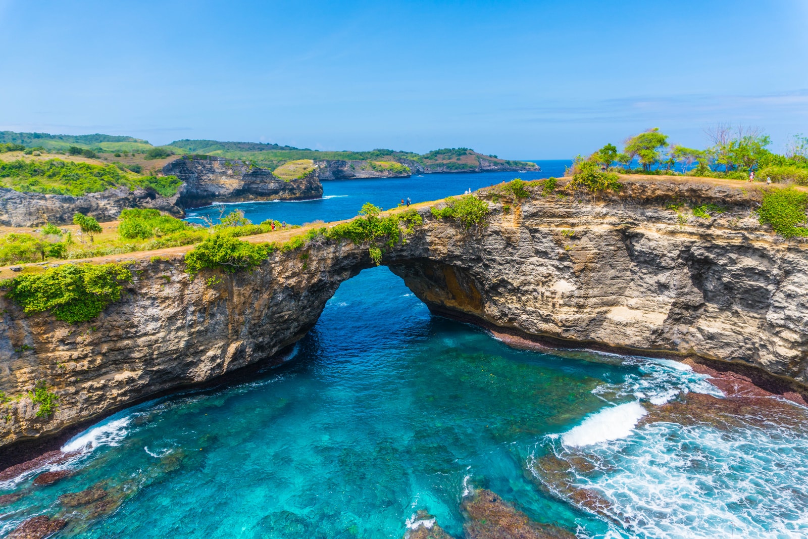 A natural rock arch over a turquoise water inlet, known as Broken Beach, under a partly cloudy sky.