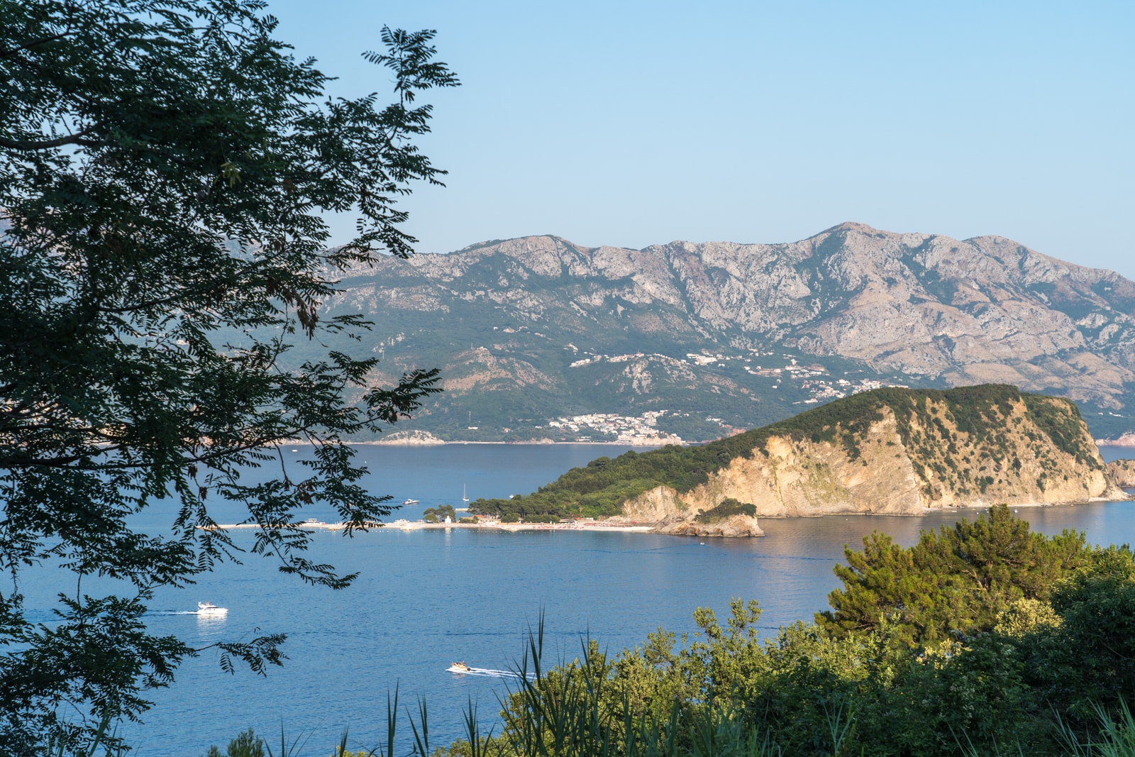 A wide view over Budva Bay shows the city on the right, the large, forested Sveti Nikola Island in the center, and lush green hills in the foreground.
