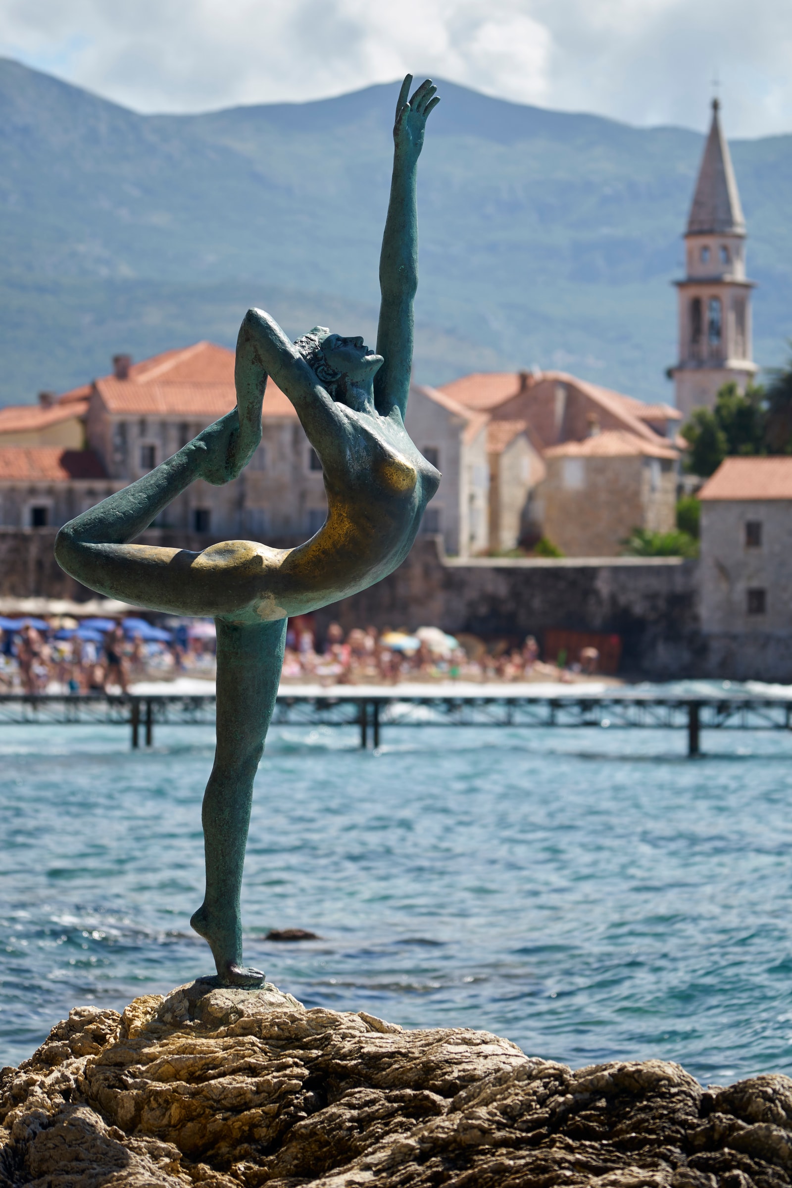 A daytime close-up shot of the bronze dancer statue shows its weathered green patina, with the stone walls of Budva's Old Town in the background.
