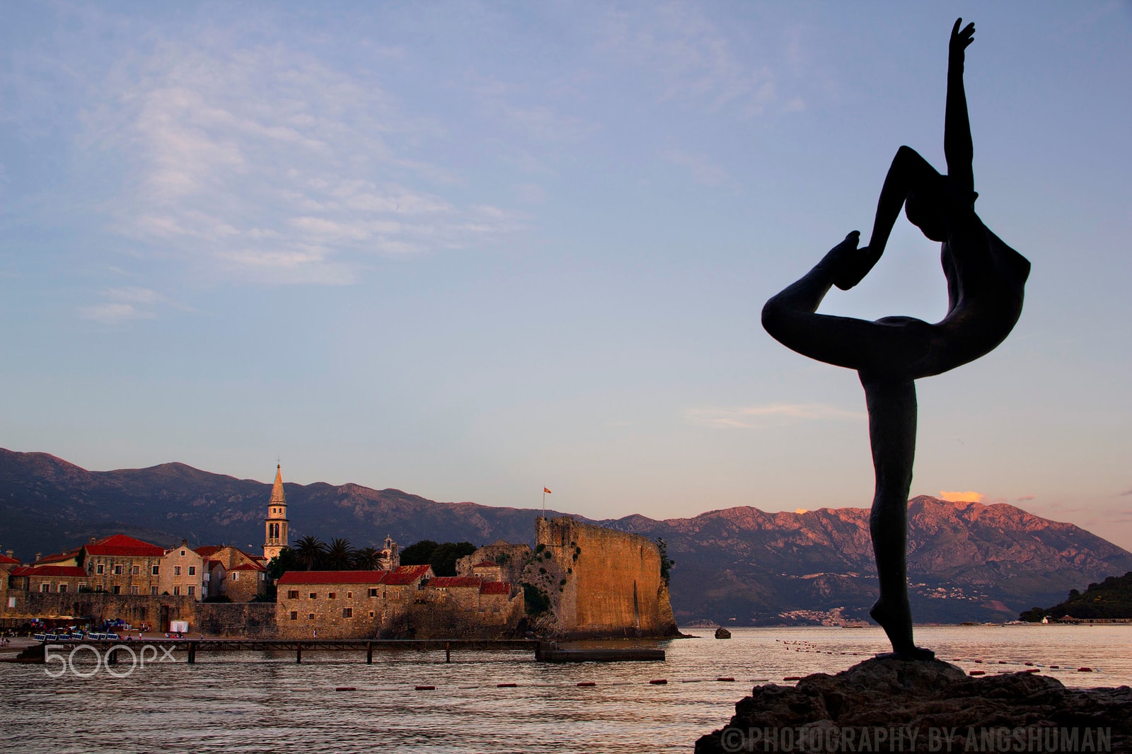 A bronze statue of a female dancer in an elegant pose stands on a coastal rock at sunset, with the historic walls of Budva's Old Town in the background.