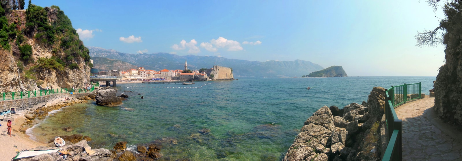 A panoramic view from a beach shows Budva's Old Town and the large, green Sveti Nikola Island in the bay.