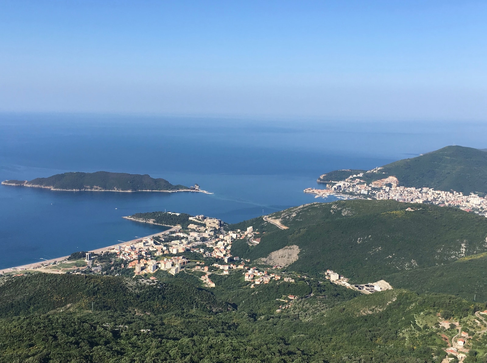 A high-angle aerial photo shows the sweeping curve of the Montenegrin coastline, with the towns of Bečići and Budva visible along the shore and Sveti Nikola island offshore.