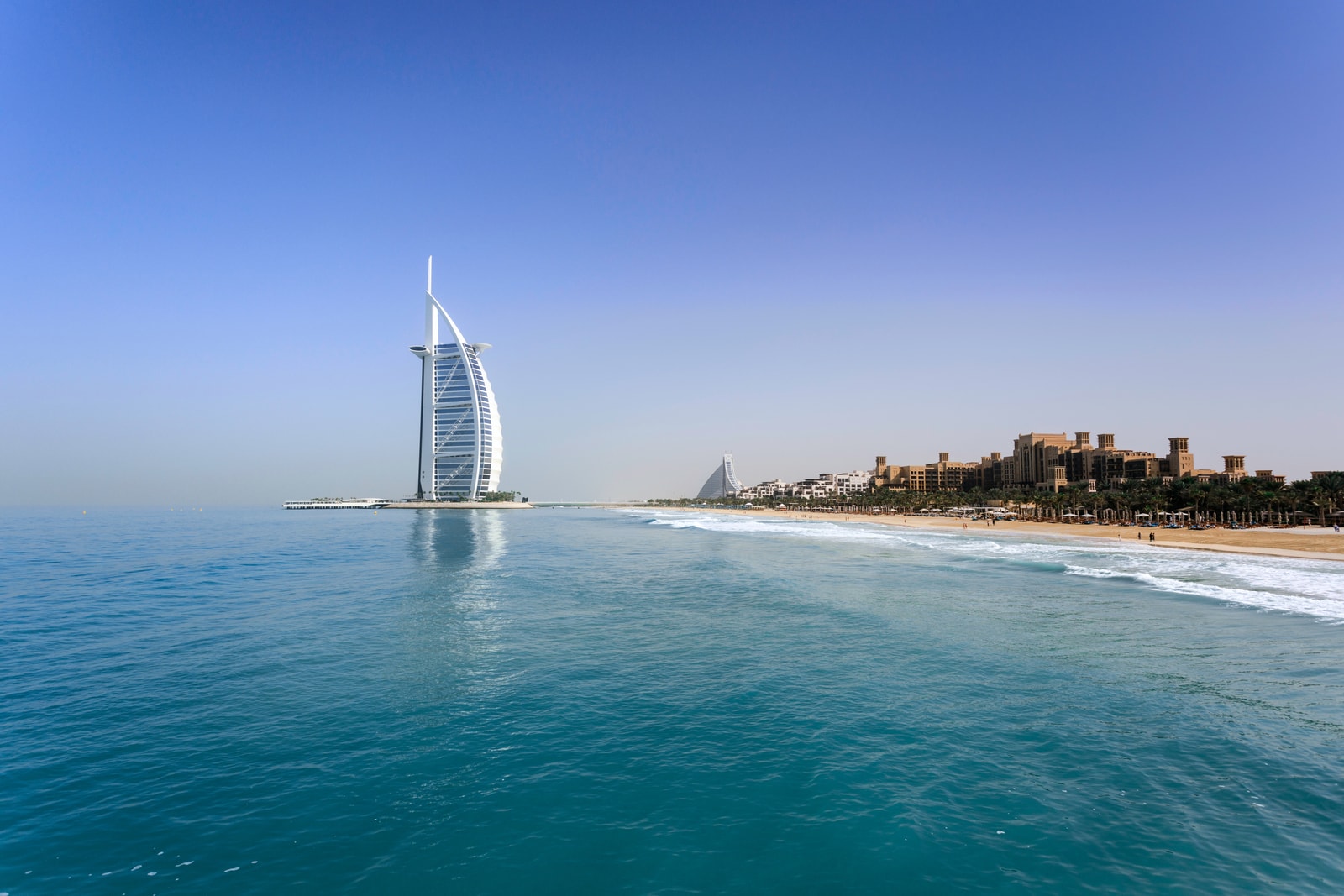 A clean, high-resolution view of the Burj Al Arab from the coast, with a curved beach in the foreground and clear blue water.