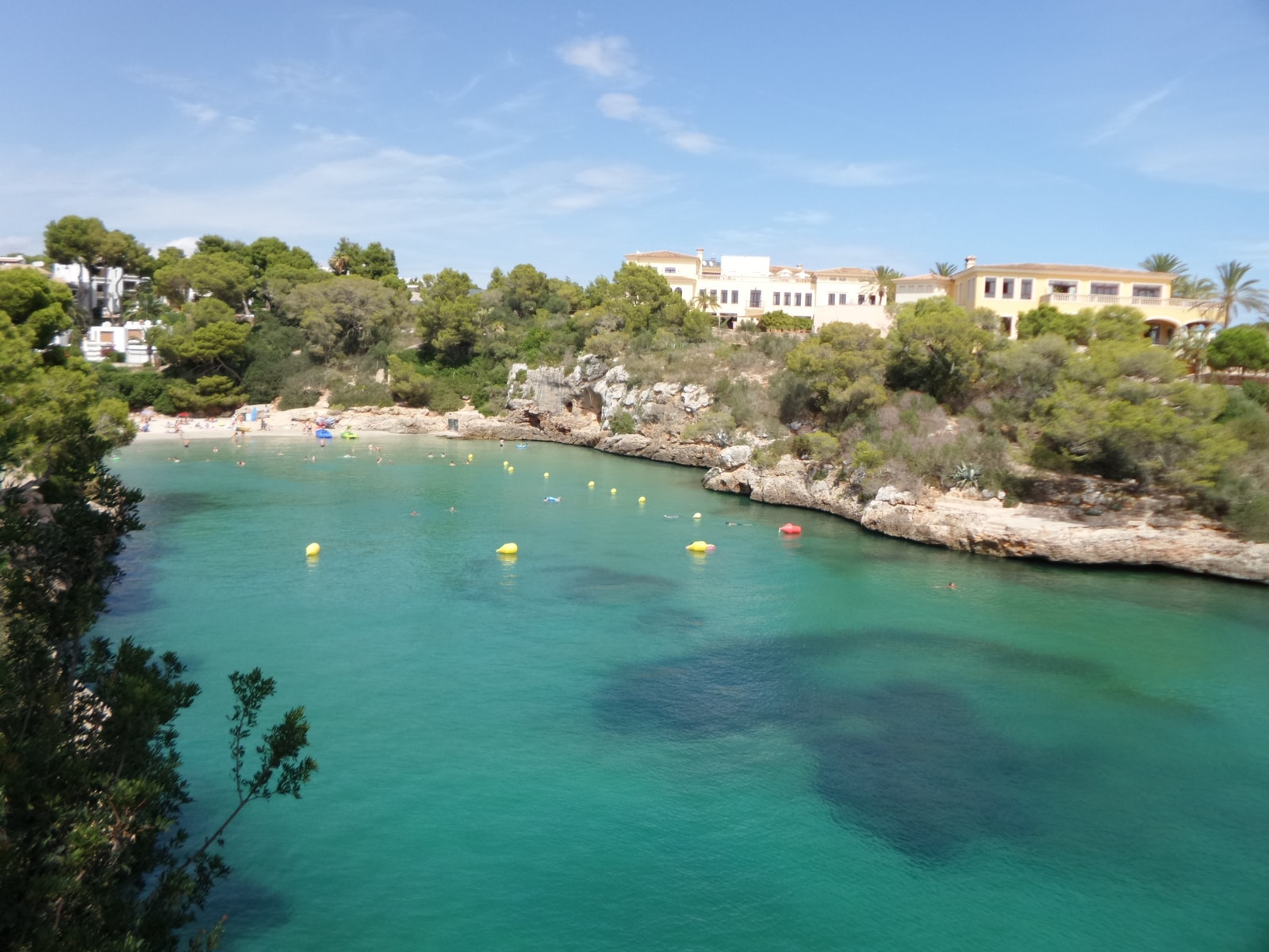 A view from the water shows the cove of Cala Ferrera, with its turquoise sea, rocky shores, and resort buildings nestled among the trees.