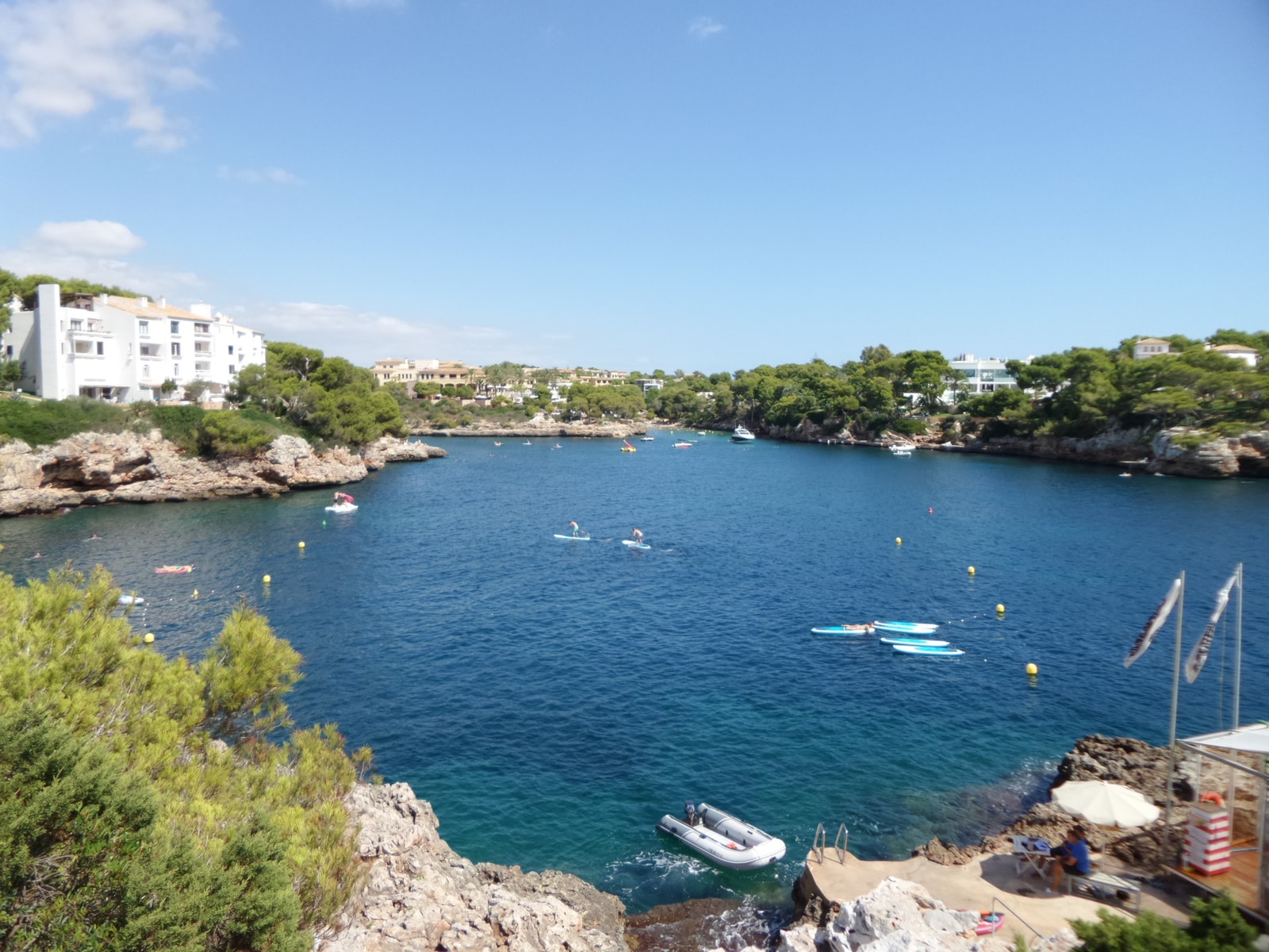An elevated view of the Cala Ferrera inlet shows swimmers and a paddleboat in the blue-green water, with rocky shores and resort buildings in the distance.