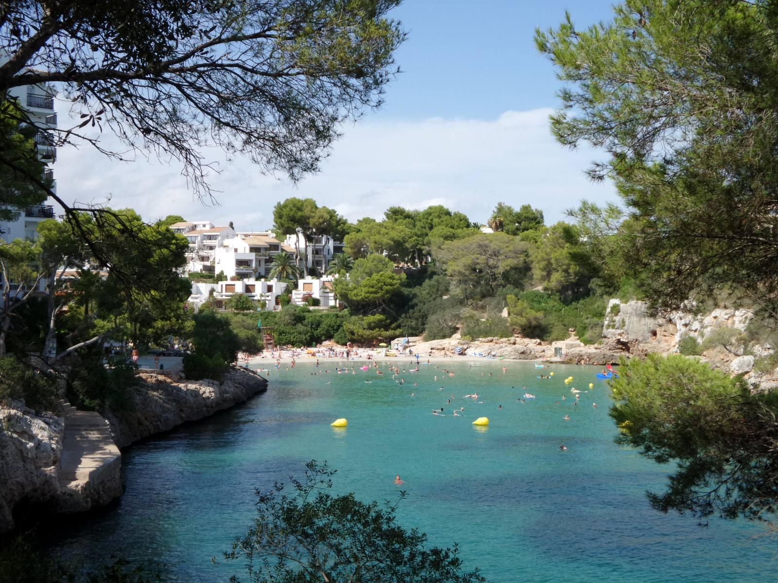 A view from a rocky walkway shows pine trees framing the turquoise water of Cala Ferrera's inner cove.