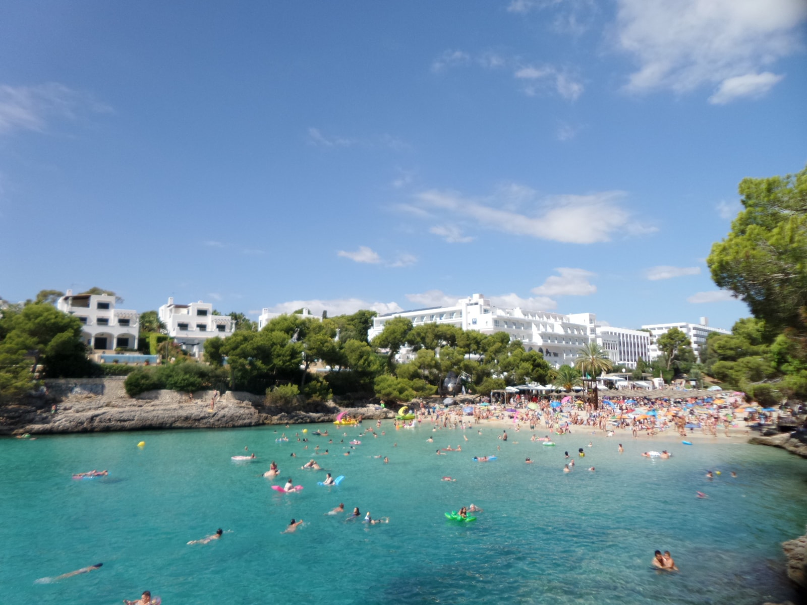 Swimmers enjoy the turquoise water of Cala Gran on a sunny day, with the sandy beach and resort buildings visible in the background.
