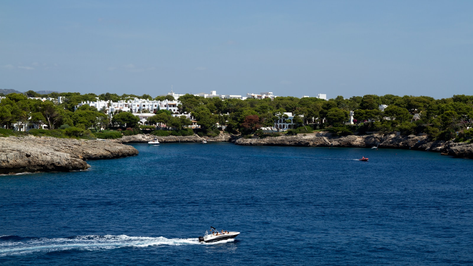 A wide view of the Cala Gran inlet shows deep blue water surrounded by rocky, pine-covered headlands and the shoreline of the resort.