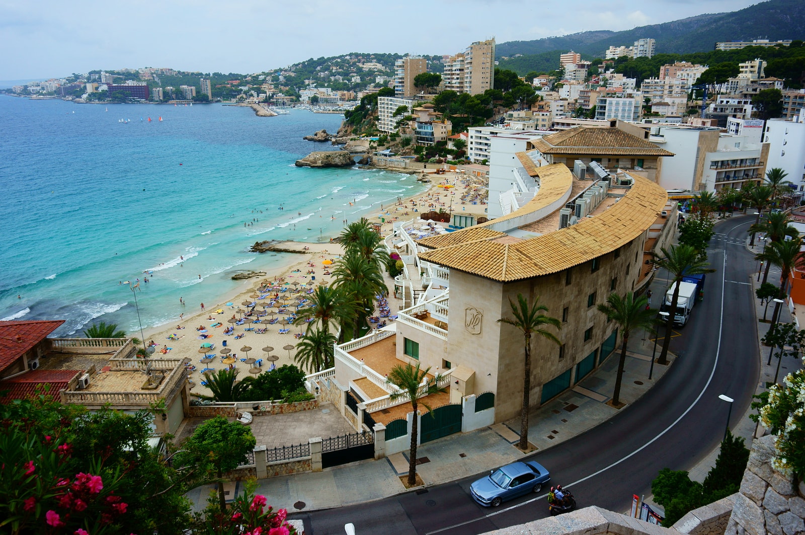 An elevated view of a sandy cove beach, with blue-green water, resort buildings, and palm trees surrounding it.