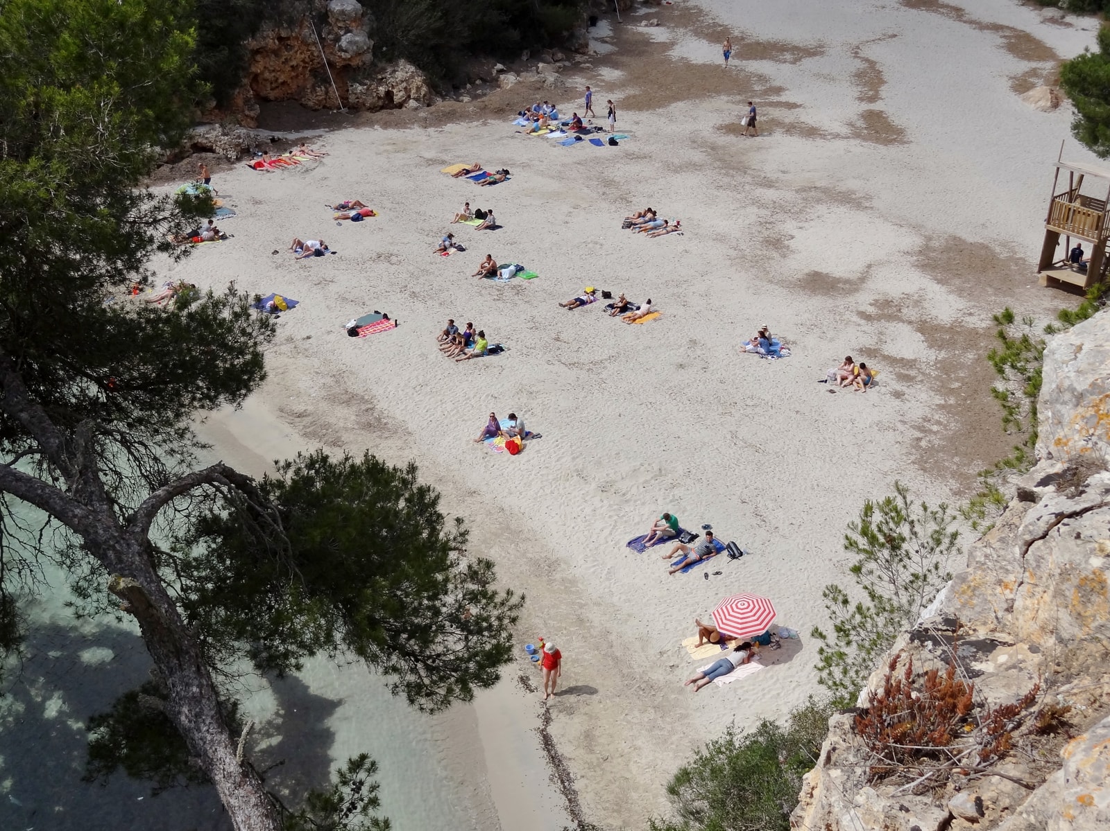 A view from above of a narrow sandy beach nestled between dramatic rocky cliffs, with turquoise water and pine trees framing the scene.