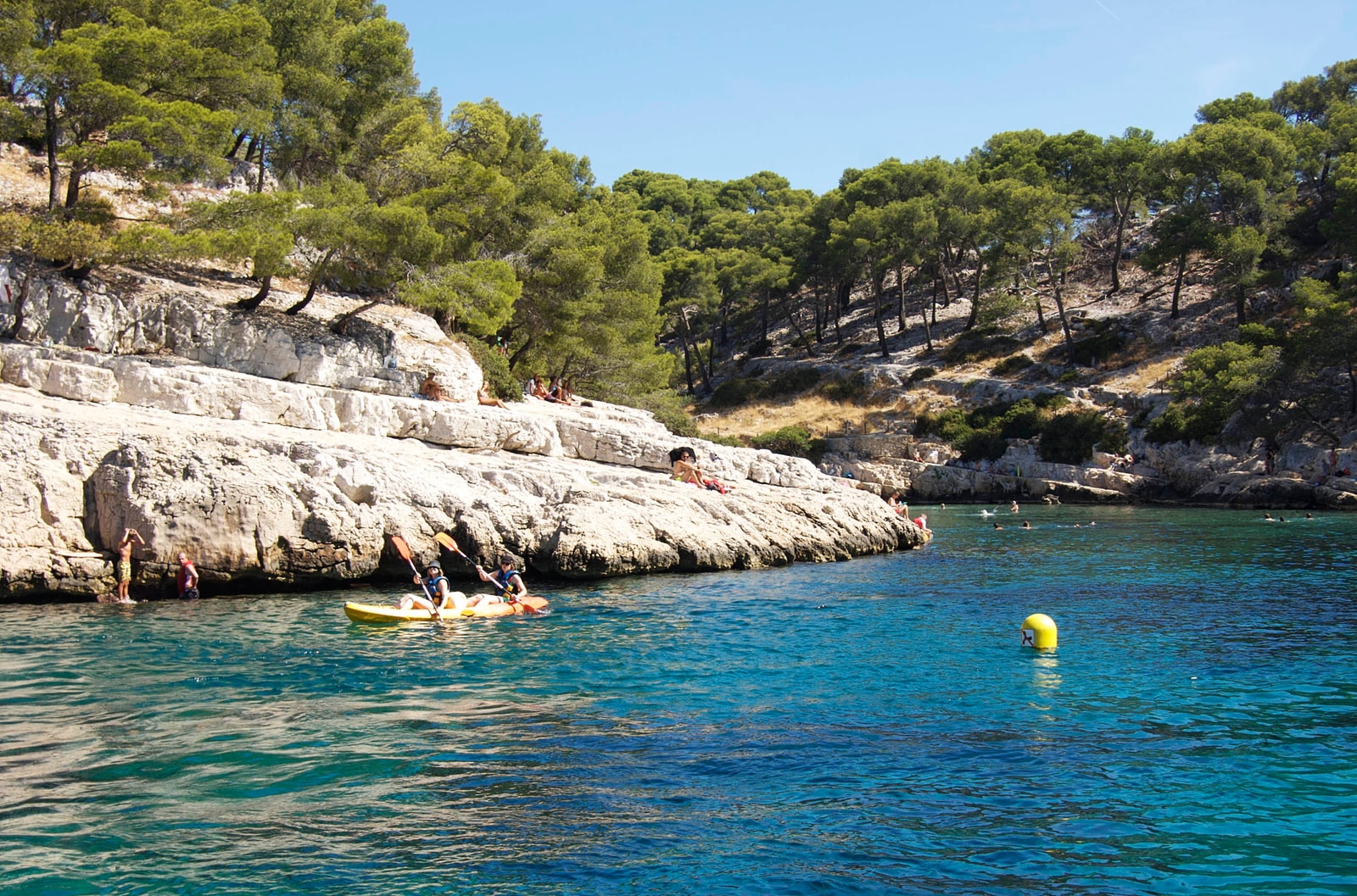 People in kayaks paddle on the bright turquoise water of Calanque de Port-Pin, with others relaxing on the white limestone ledges under pine trees.