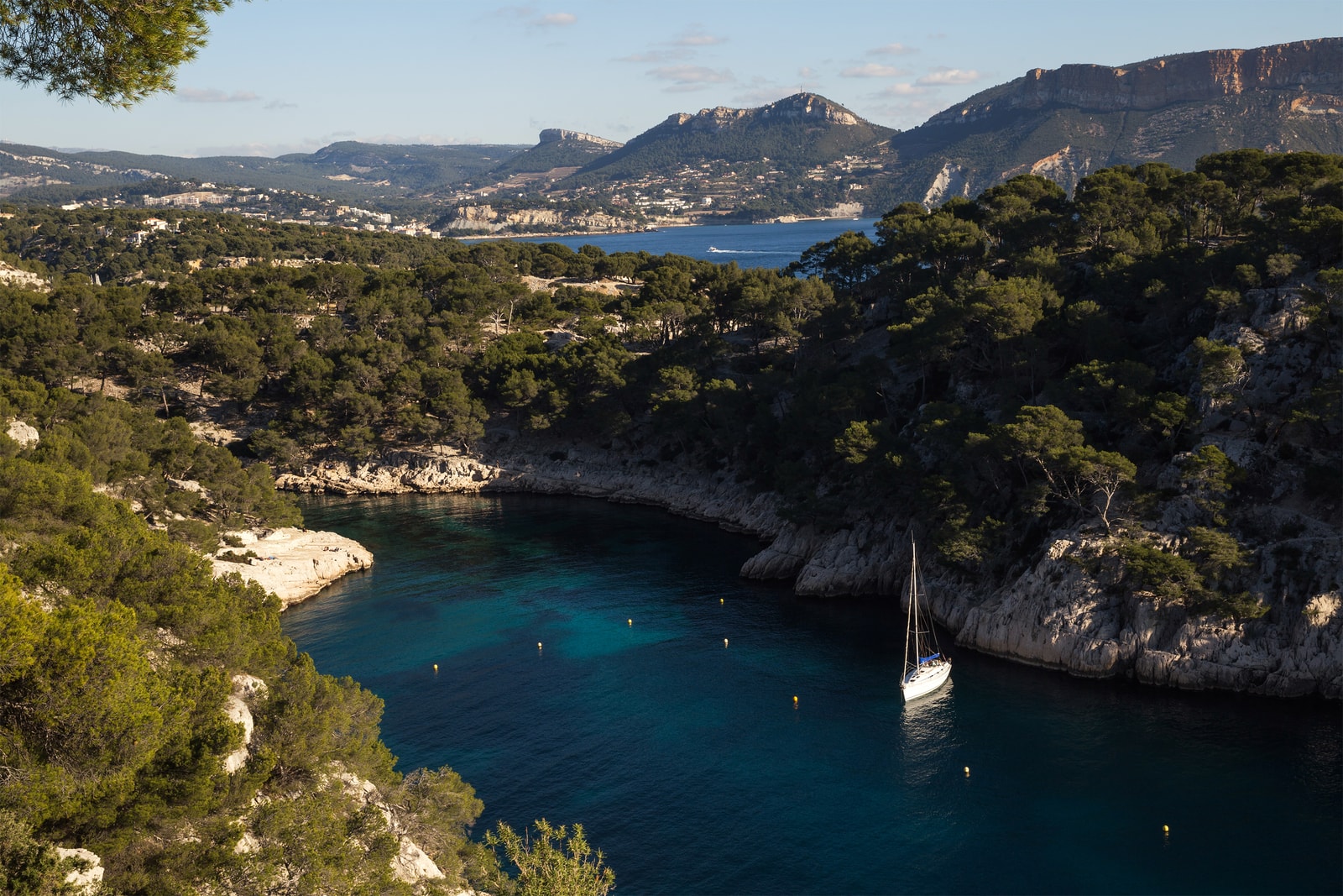 A single white sailboat is anchored in the calm, deep blue water of Calanque de Port-Pin, surrounded by steep, pine-covered slopes.