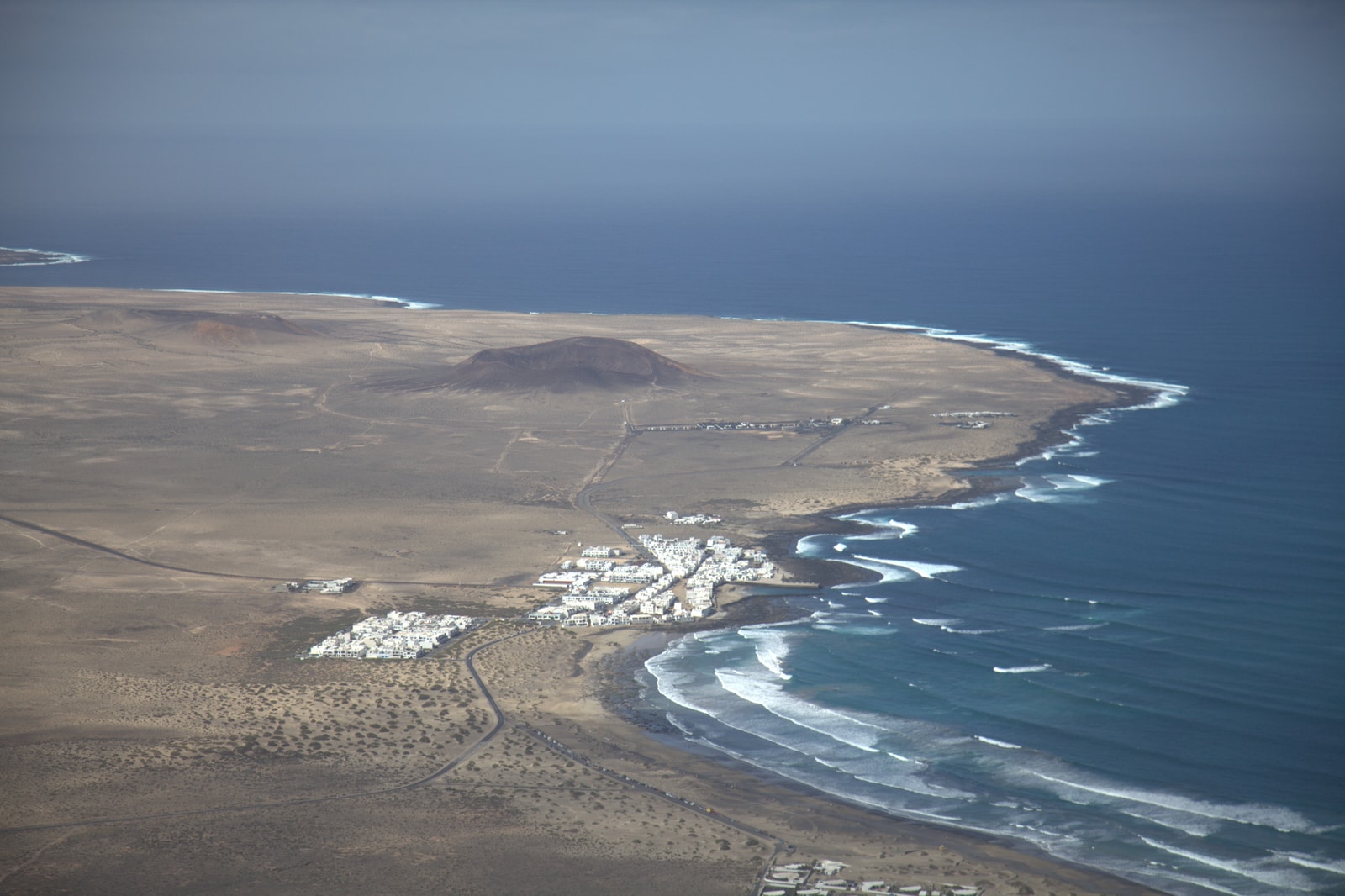 An aerial shot shows the town of Caleta de Famara on a wide, sandy coastline with waves breaking offshore.