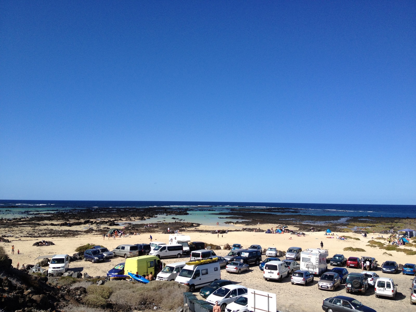 A beach with white sand and shallow turquoise water is dotted with dark volcanic rocks.