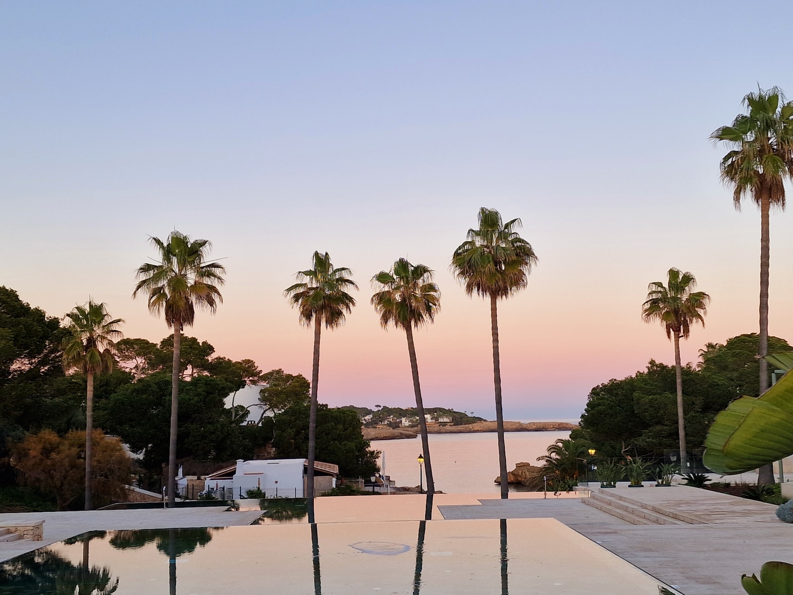 Palm trees are reflected in the calm water of the Caló d'es Pou inlet at dusk, with a pastel-colored sky overhead.