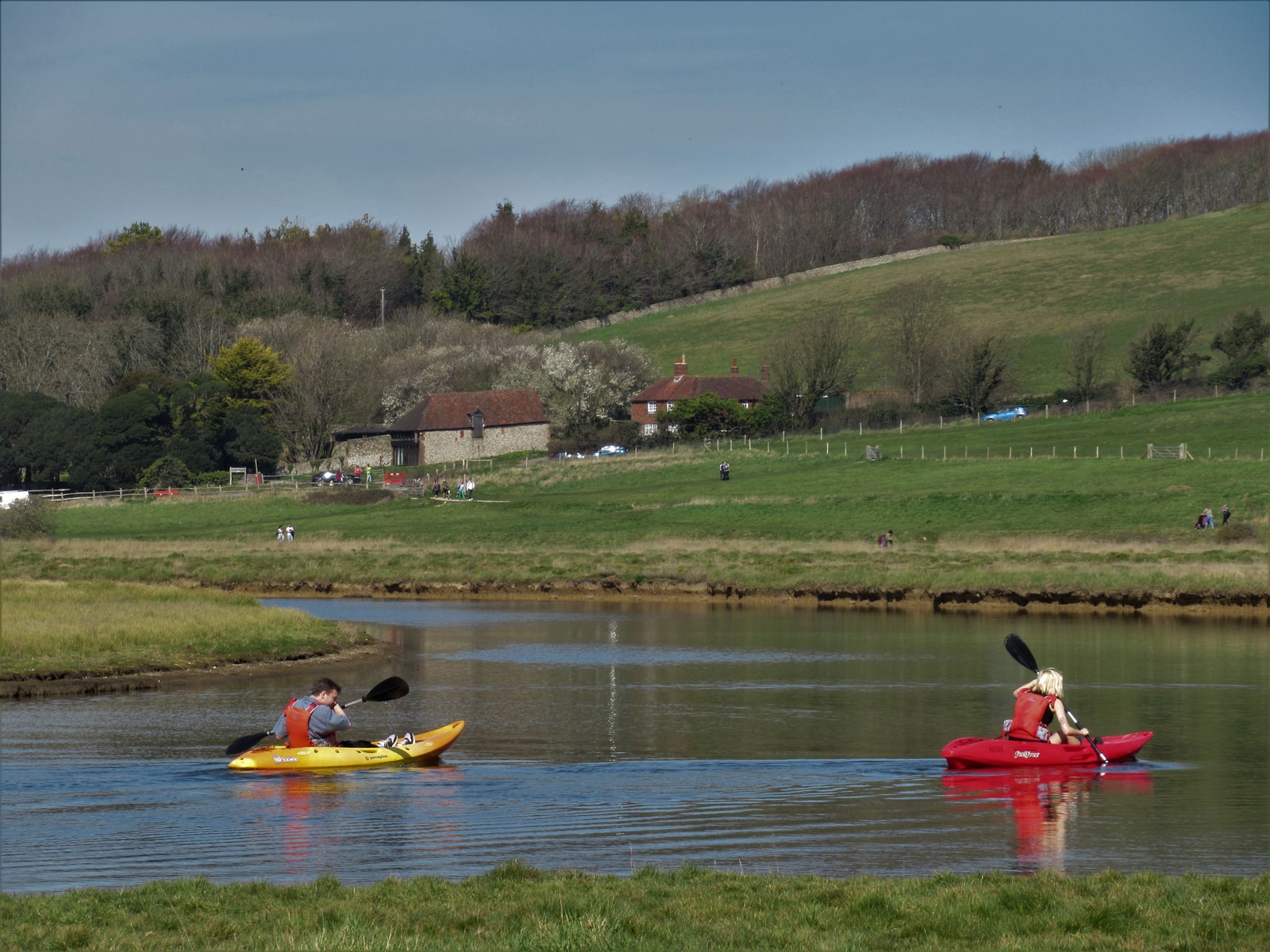 Two people in kayaks paddle down the calm, green waters of the Cuckmere River within Seven Sisters Country Park.