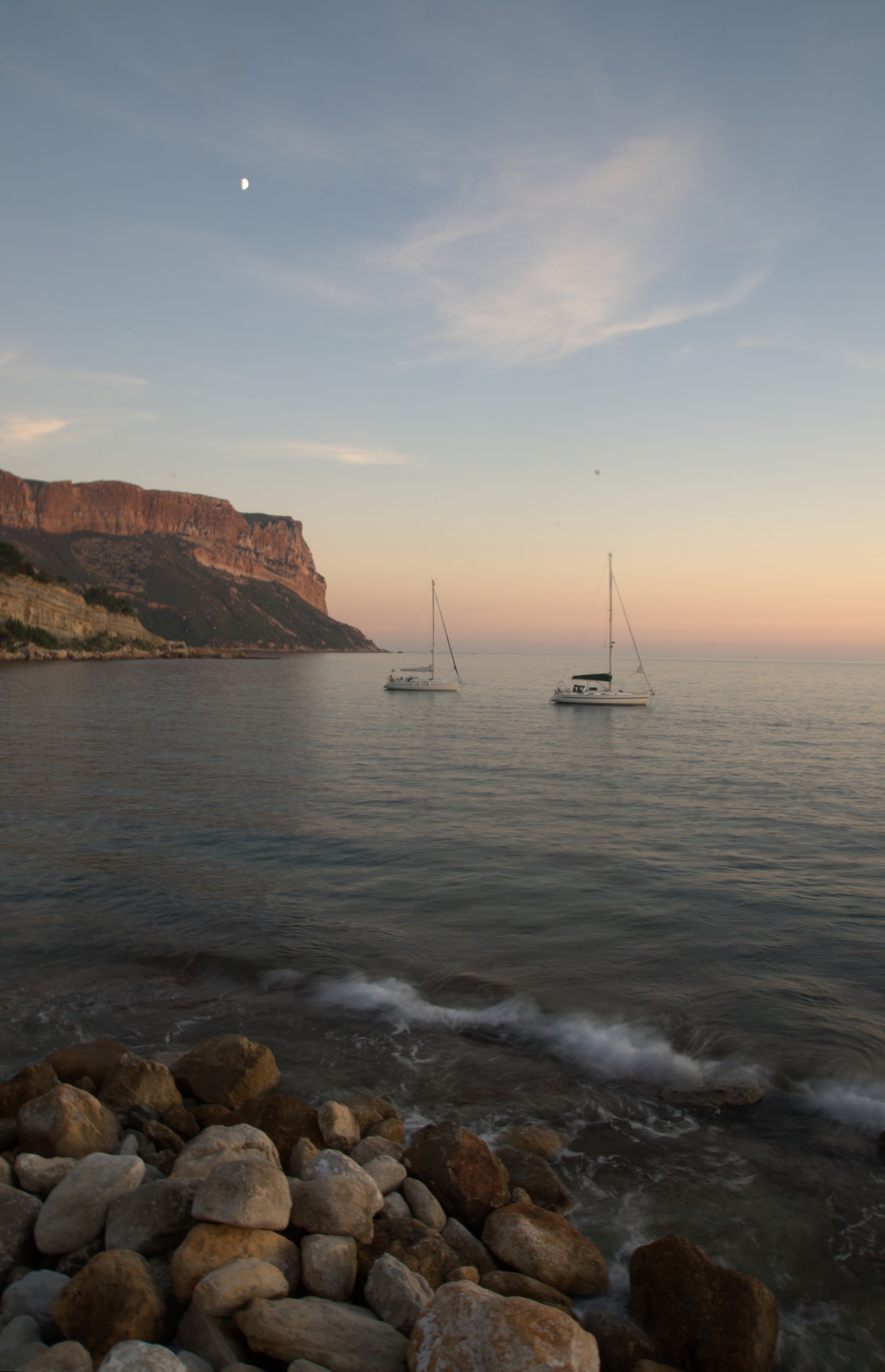 Smooth beach pebbles in the foreground with gentle waves under a twilight sky tinted orange behind the Cap Canaille sea cliffs.
