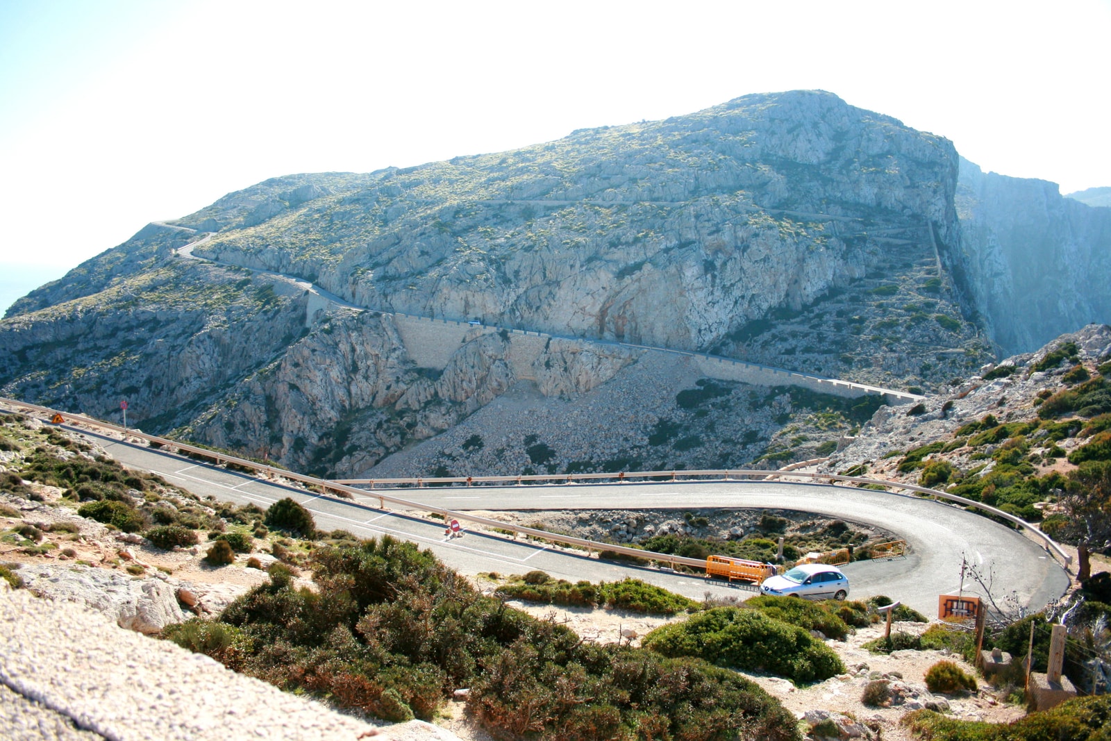 A winding asphalt road cuts through a rugged, mountainous landscape with sparse vegetation under a clear blue sky.