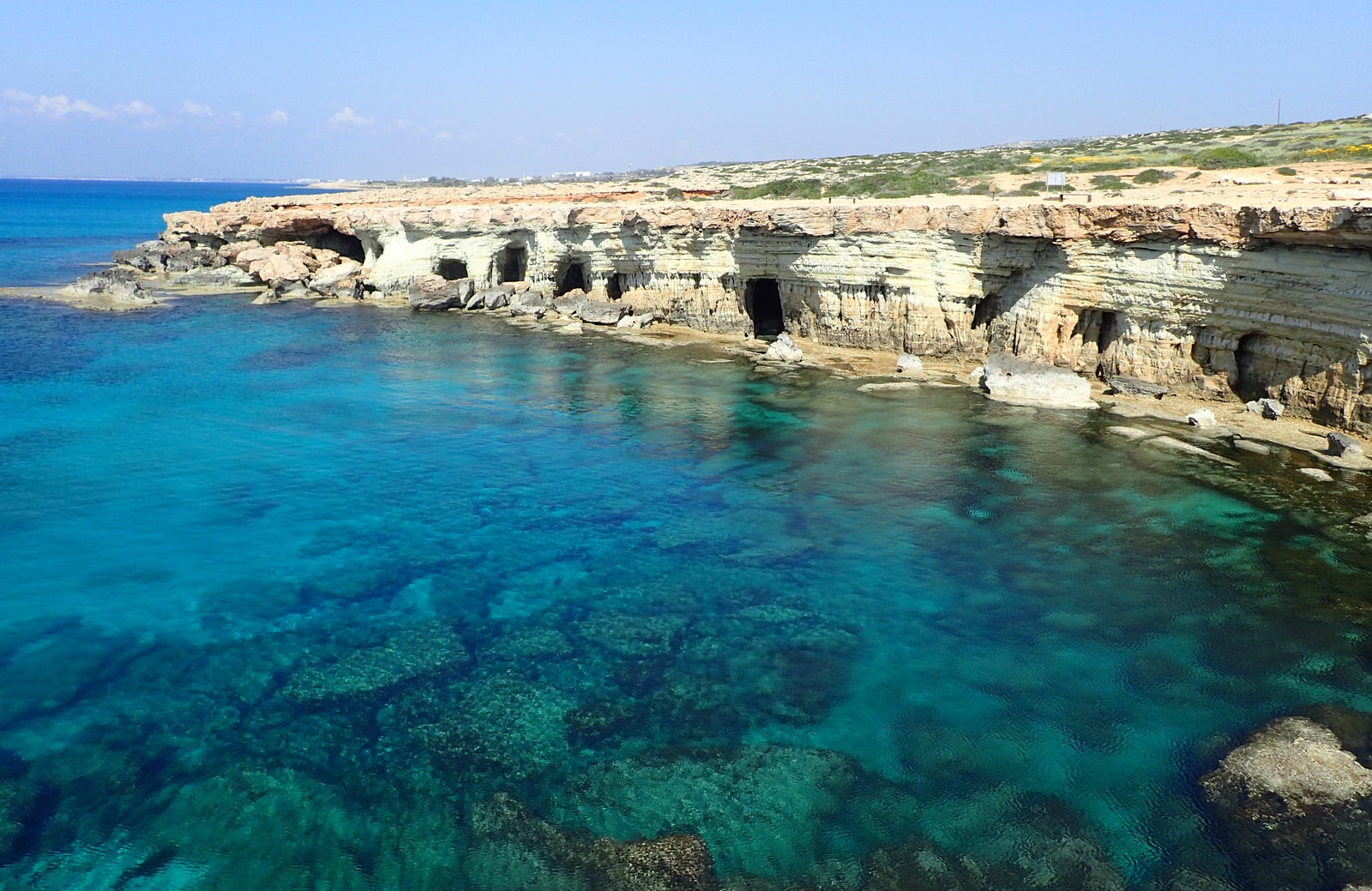 The rugged limestone coastline of Cape Greco, showing clear turquoise water in a small inlet and sea caves in the distance.