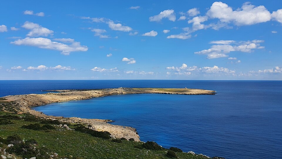 A panoramic view of the rugged Cape Greco coastline, with scrubby vegetation in the foreground and the turquoise sea stretching to the horizon under a clear blue sky.