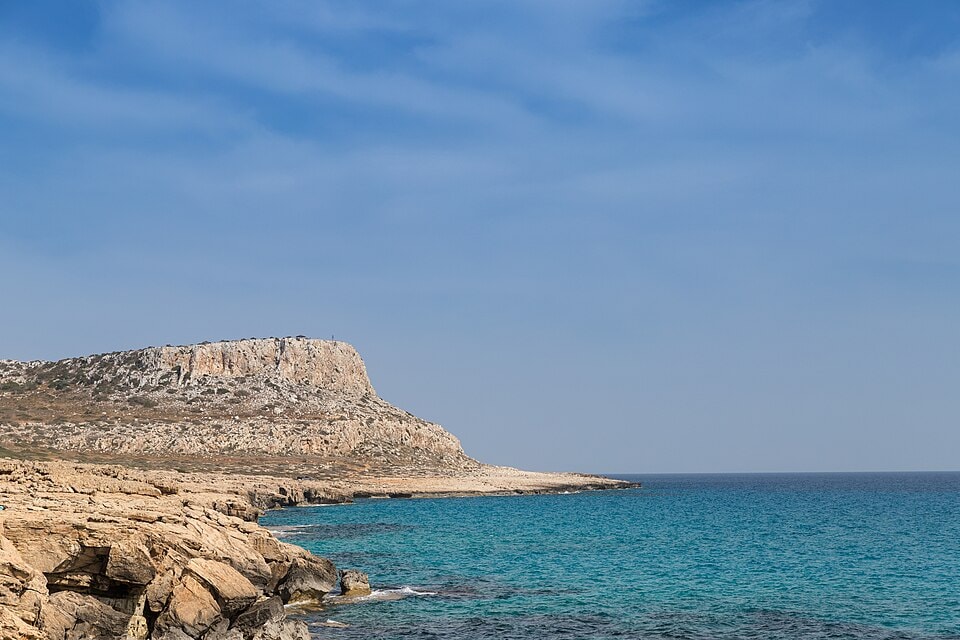 The rugged limestone cliffs and deep blue water of Cape Greco, Cyprus, under a clear sky.