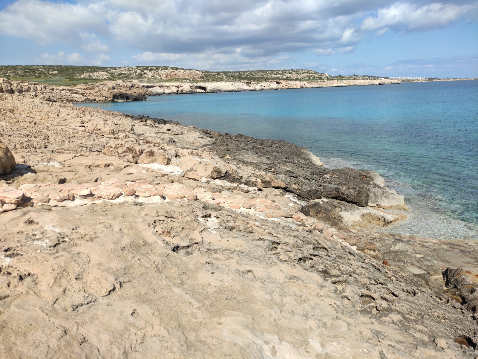 The rugged, layered cliffs of Cape Greco meet the turquoise Mediterranean Sea under a clear blue sky.
