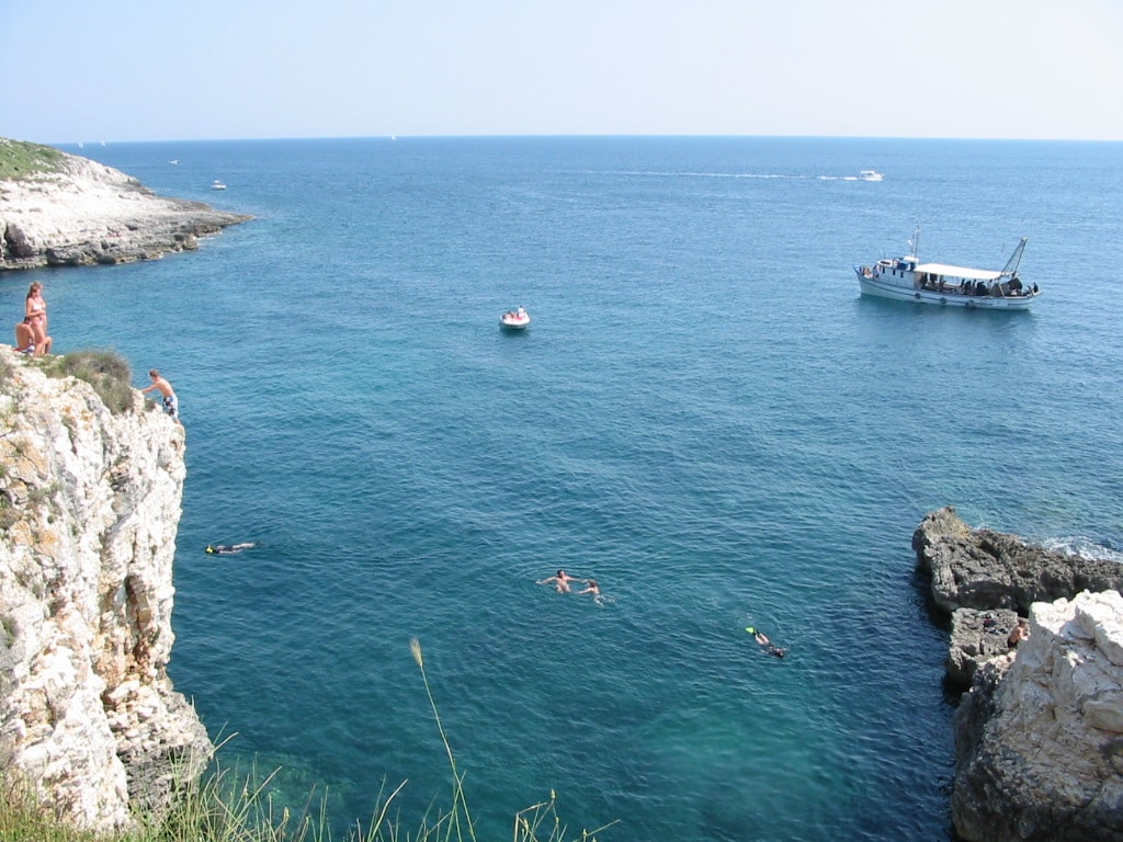 People swimming and cliff jumping from large stone slabs into the clear blue water at Cape Kamenjak on a sunny day.