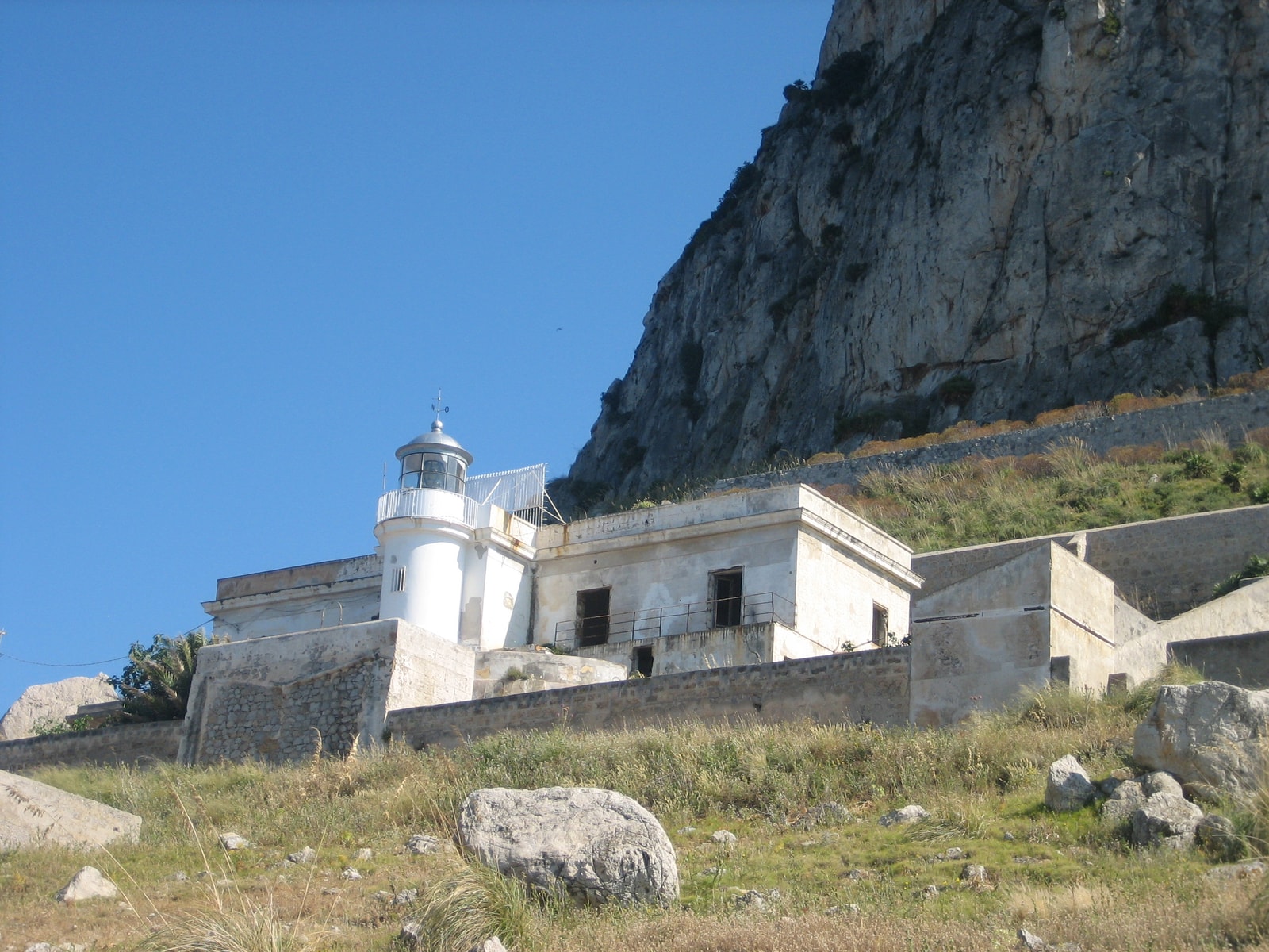 The Faro di Capo Gallo lighthouse, a white structure situated at the base of the towering, rugged limestone cliffs of Monte Gallo.