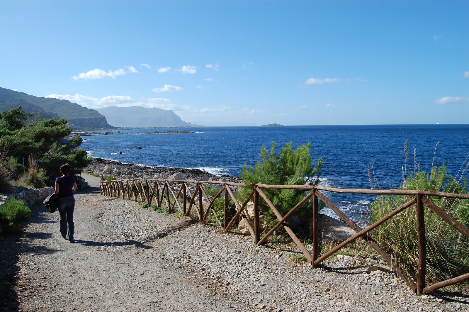 A view of a hiking path within the Capo Gallo Nature Reserve, with a rustic wooden fence along the trail and the sea in the distance.