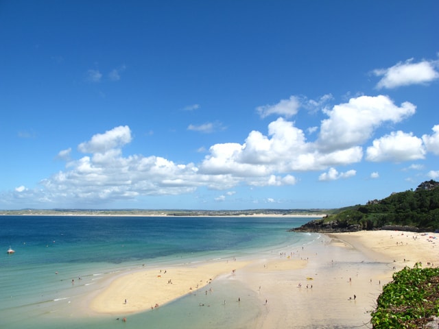 A panoramic view of Carbis Bay beach shows its wide crescent of golden sand meeting calm, turquoise water under a blue sky.