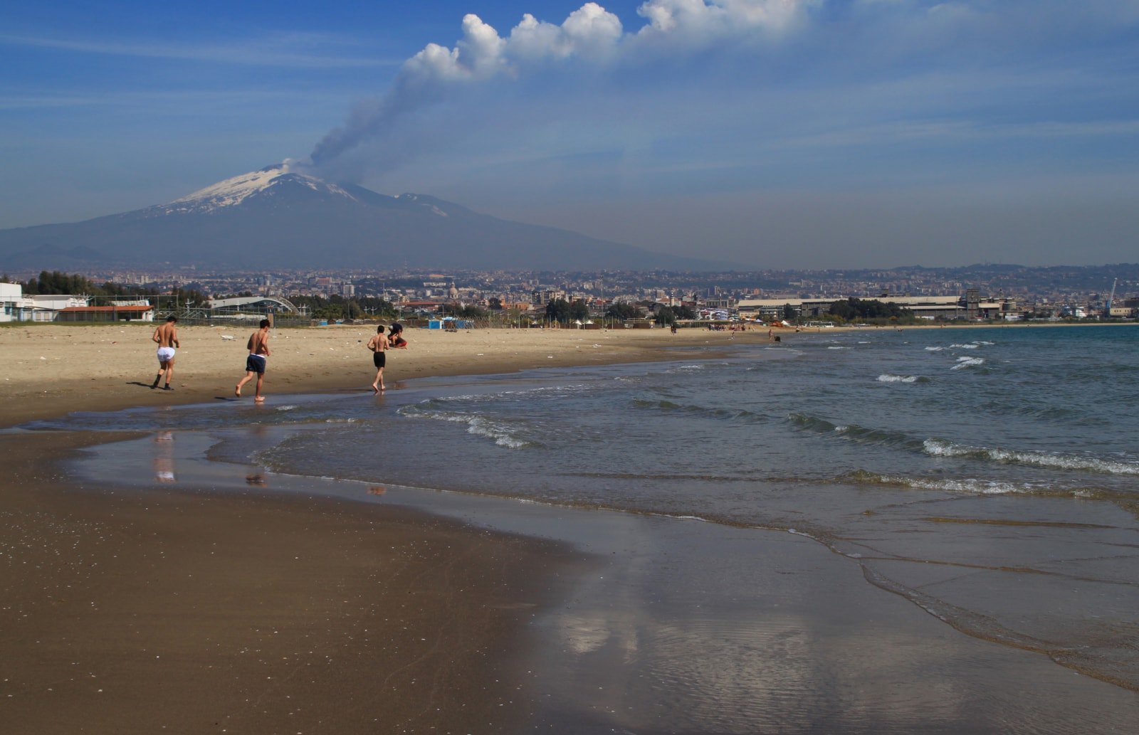 Catania's long sandy shore, La Playa, with joggers in the foreground and a smoking, snow-dusted Mount Etna rising in the background.