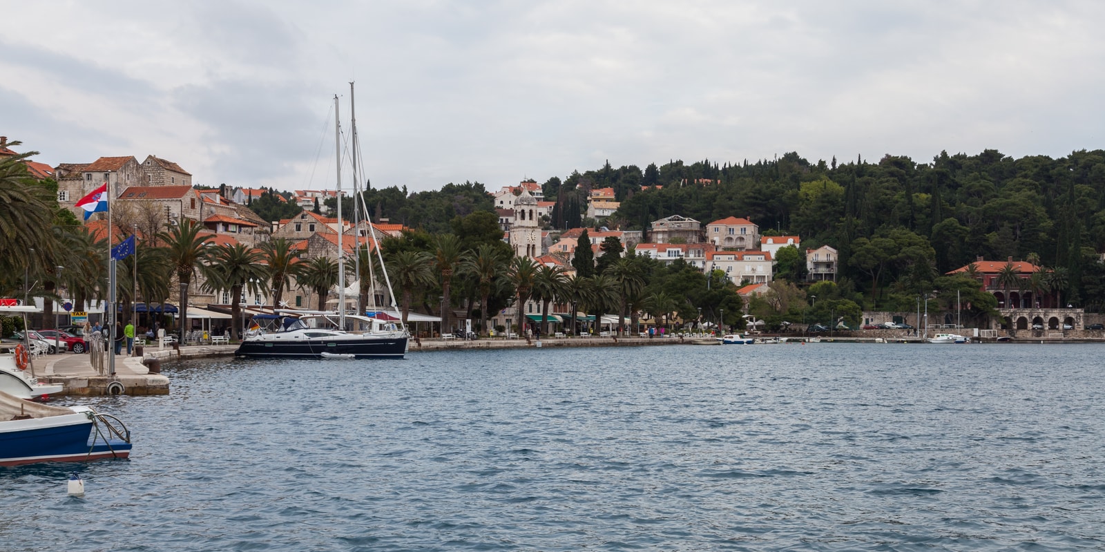 A wide-angle shot of a waterfront promenade with palm trees, boats in the harbor, and red-roofed buildings against a green hillside.