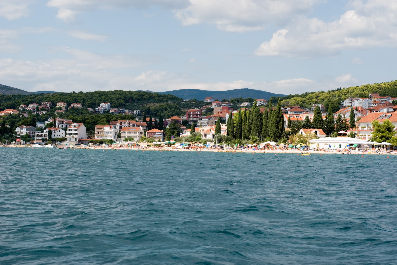A panoramic view of the Čiovo coastline, showing waterfront settlements, wooded hills, and the calm Adriatic Sea.