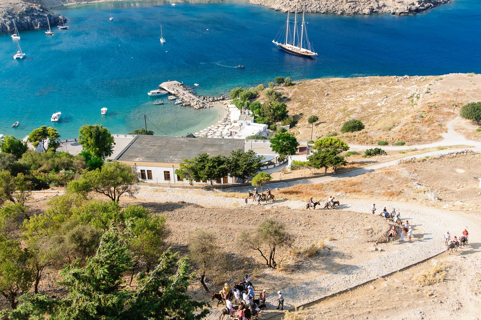 Visitors walk up a path on a dry, rocky hillside, with the bright blue water of the bay visible below.