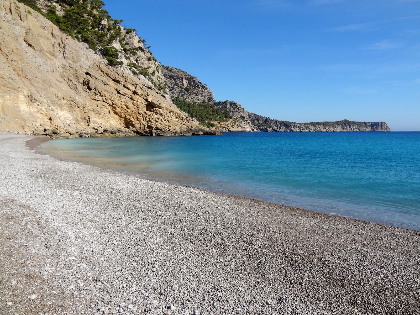 A view of a coarse pebble beach with clear, turquoise water under towering limestone cliffs.